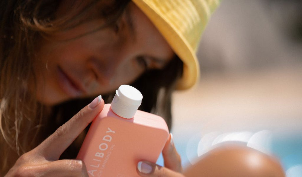 Frau mit gelbem Hut betrachtet konzentriert eine rosafarbene Lotion-Flasche mit der Aufschrift ‚BALIBODY‘ bei Sonnenlicht am Pool.