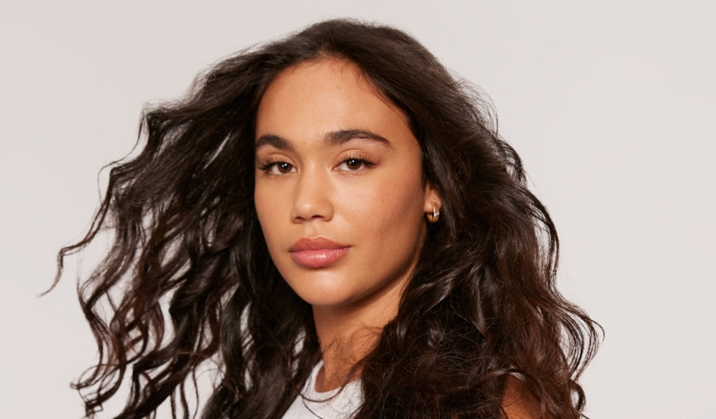 Frau mit langen, dunklen Locken blickt selbstbewusst in die Kamera, trägt schlichte Ohrringe und ein weißes Oberteil vor neutralem Hintergrund.