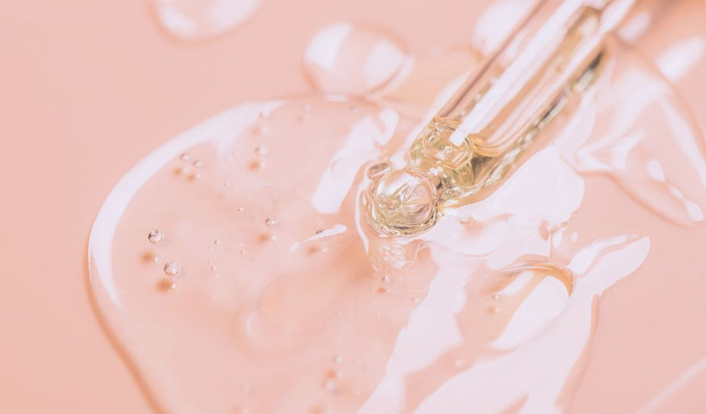 Nahaufnahme einer Pipette mit durchsichtigem Serum auf rosafarbenem Untergrund, mit glänzender Textur und kleinen Bläschen.