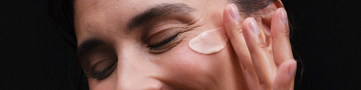 Eine Person trägt mit geschlossenen Augen lächelnd Creme auf ihre Wange auf, vor einem dunklen Hintergrund.