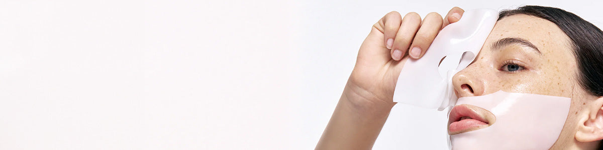 Frau, die vor einem schlichten weißen Hintergrund eine weiße Tuchmaske von ihrem Gesicht entfernt.