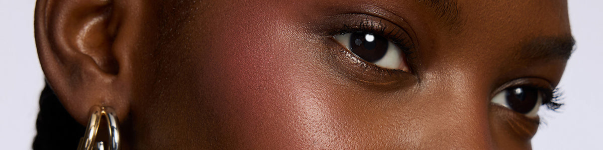 Nahaufnahme der Wange einer Person, welche mit Blush hervorgehoben wird. Sie zeigt glatte Haut, dezentes Make-up und einen goldenen Ohrring.