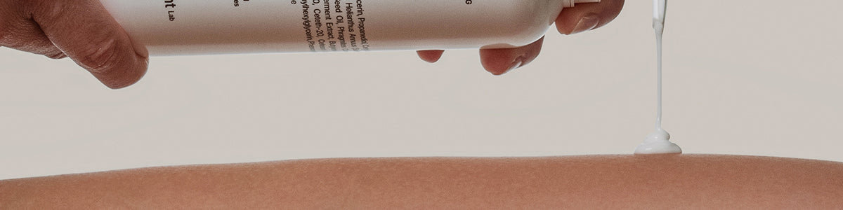 Vor einem neutralen Hintergrund gießt eine Hand weiße Lotion aus einer Flasche auf den Unterarm einer Person.