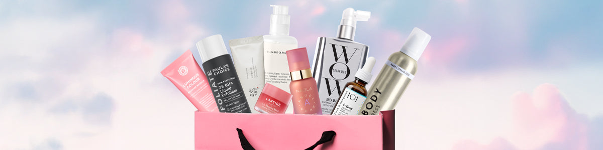 Pinke Geschenktasche mit verschiedenen Beauty-Produkten vor pastellfarbenem Himmel.