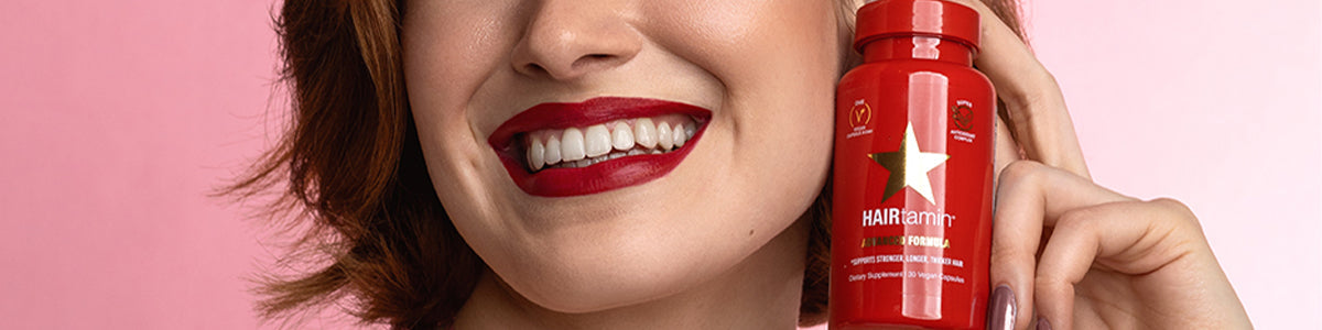Lächelnde Frau mit rotem Lippenstift, die vor einem rosa Hintergrund einen roten Behälter mit der Aufschrift „Hairtamin“ hält.