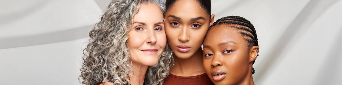 Drei Frauen mit unterschiedlichen Hauttönen stehen eng beieinander und lächeln sanft in die Kamera.