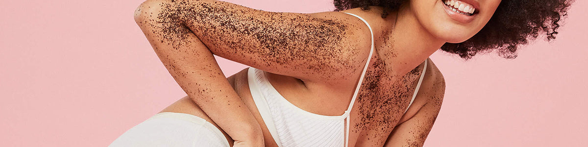 Lächelnde Frau in weißer Unterwäsche mit Kaffeepeeling auf Schulter und Arm vor rosa Hintergrund.