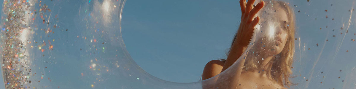 Frau blickt durch einen transparenten, glitzernden Schwimmring in die Kamera vor blauem Himmel.