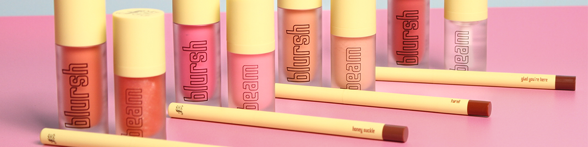 Bunte Blursh- und Beam-Lipglosse sowie Lipliner, angeordnet auf einer rosa Oberfläche.