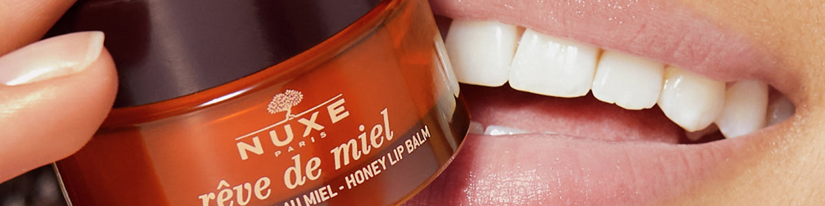 Nahaufnahme einer lächelnden Person, die ein Glas Nuxe Rêve de Miel Honig-Lippenbalsam an die Lippen hält.