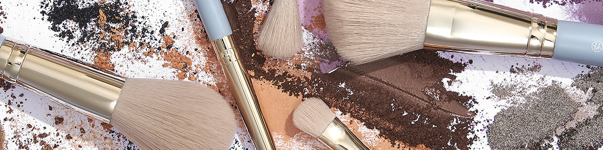 Make-up-Pinsel mit goldenen Griffen liegen auf verstreutem Puder, Lidschatten und Rouge in verschiedenen Farben.