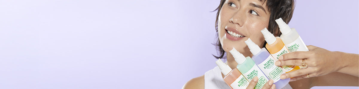 Eine Frau hält fünf farbige Sprayflaschen in der Hand und lächelt, vor einem hellvioletten Hintergrund.