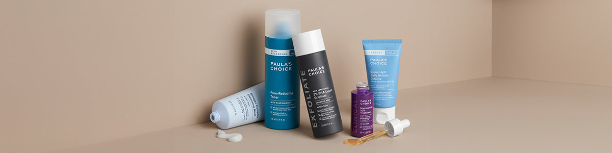 Fünf Hautpflegeprodukte von Paula’s Choice, angeordnet auf einer beigen Oberfläche, mit einer Pipette und kleinen Tropfen Lotion in der Nähe.