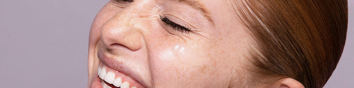 Lachende Frau mit Sommersprossen und geschlossenen Augen; ein transparenter Tropfen Hautpflegeprodukt ist unter dem rechten Auge aufgetragen.