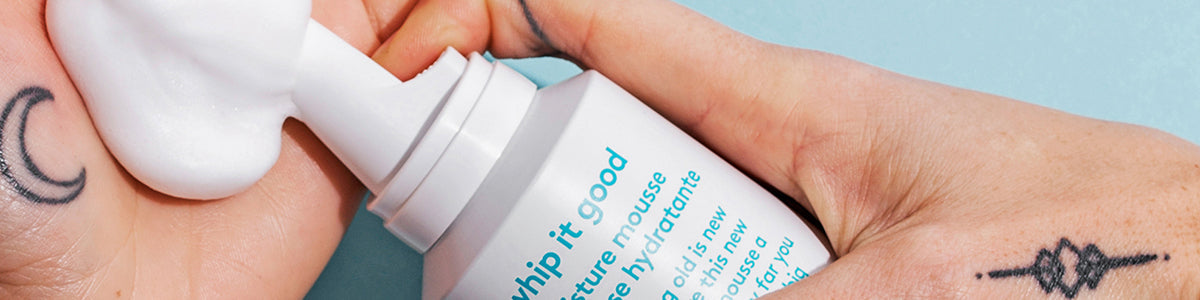 Hand mit Tätowierungen, die vor blauem Hintergrund weißen Schaum aus einer Flasche mit der Aufschrift „Whip It Good Moisture Mousse“ ausgibt.