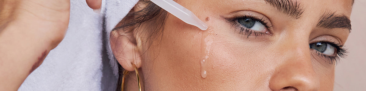 Frau mit Handtuch um den Kopf trägt mithilfe einer Pipette ein flüssiges Serum auf die Haut unterhalb des Auges auf, ein Tropfen läuft über die Wange.