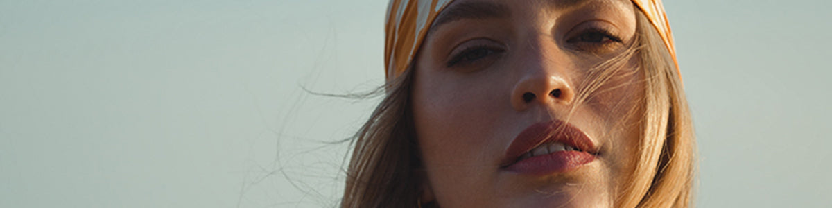 Frau mit langen Haaren und gelbem Kopftuch, die vor einem blassen Himmelshintergrund in die Kamera blickt.