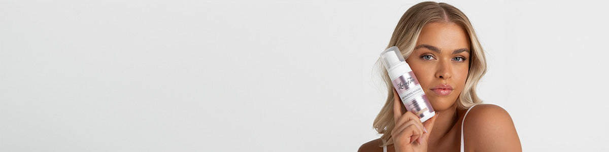 Frau mit blonden Haaren hält eine weiße Flasche mit Selbstbräunungsschaum vor ihr Gesicht und blickt in die Kamera.