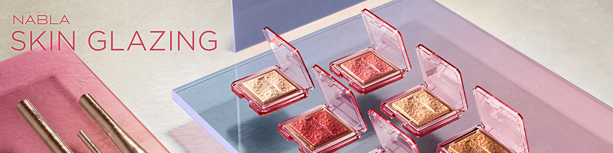 Nabla Skin Glazing Highlighter in offenen, roten Kompaktboxen auf Glasplatten, daneben goldene Stifte, in einem modernen Setting arrangiert.