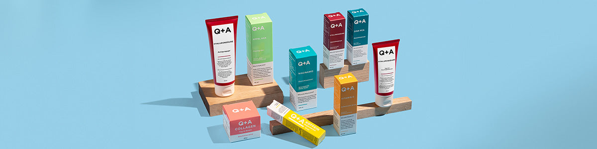 Bunte Hautpflegeprodukte der Marke Q+A in Tuben und passenden Boxen, dekorativ auf Holzblöcken vor hellblauem Hintergrund arrangiert.