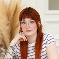 Porträt von Pauline mit roten Haaren, Brille mit goldenem Rahmen und rot weiß gestreiftem Oberteil vor hellem Hintergrund.