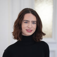 Porträt von Karolin mit dunkelbraunen Haaren, blauen Augen und schwarzem Oberteil vor hellem Hintergrund.