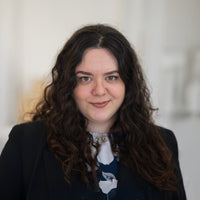 Porträt von Esra mit dunkelbraunen Locken, braunen Augen und schwarzem Oberteil vor hellem Hintergrund.