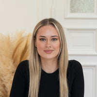 Porträt von Sara mit blonden Haaren und schwarzem Oberteil vor hellem Hintergrund.