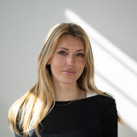 Porträt von Paula mit blonden Haaren, blauen Augen und schwarzem Oberteil vor hellem Hintergrund.