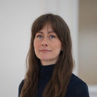 Porträt von Sylvia mit braunen Haaren, blauen Augen und schwarzem Oberteil vor hellem Hintergrund.