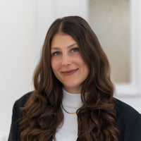 Porträt von Carina mit braunen Haaren, blauen Augen und schwarzem Blazer vor hellem Hintergrund.