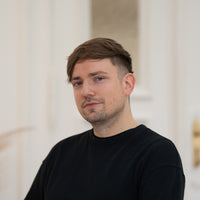Porträt von Hannes mit braunen Haaren, leichtem Bartansatz und schwarzem Oberteil vor hellem Hintergrund.
