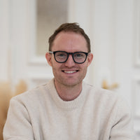 Porträt von Philipp mit braunem Haar, schwarzer Brille und beigem Oberteil vor hellem Hintergrund.
