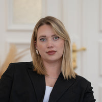 Porträt von Mara mit blonden Haaren, grünen Augen und schwarzem Blazer vor hellem Hintergrund.