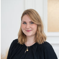 Porträt von Nora mit blonden Haaren, blauen Augen und schwarzem Oberteil vor hellem Hintergrund.
