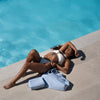 Eine Frau im weißen Bikini entspannt am Pool in der Sonne, neben sich die beiden PURISH Taschen.