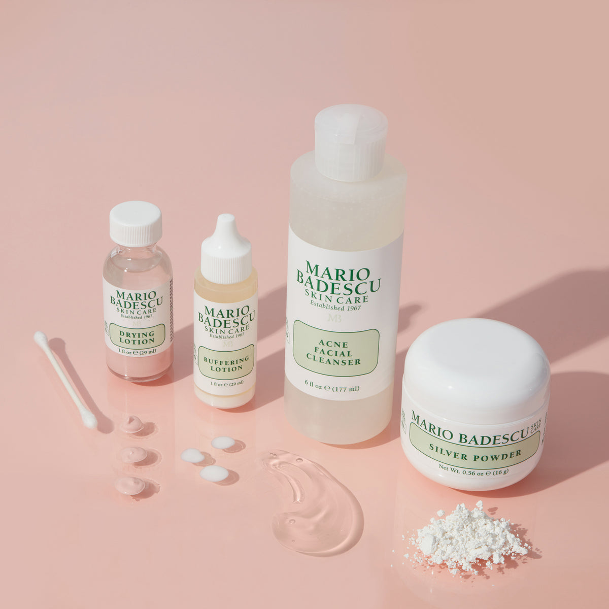 Vier Mario-Badescu-Produkte für unreine Haut auf rosa Hintergrund, mit Texturen und Wattestäbchen dekorativ arrangiert.