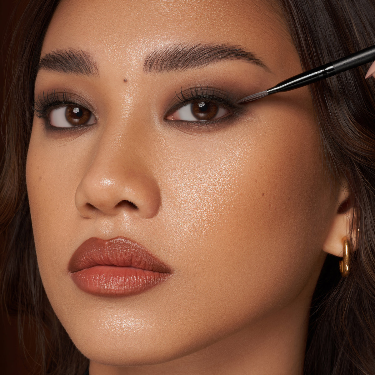 Eine Frau tmit dunklem Haar und hellem Teint trägt mit einem Make-up-Pinsel dunklen Lidschatten aus der Groundwork Palette – Defining Neutrals auf.