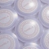 Mehrere NABLA-Puderdosen mit der Aufschrift ‚Close-Up Blotting Pressed Powder‘ in fliederfarbener Verpackung von oben fotografiert.
