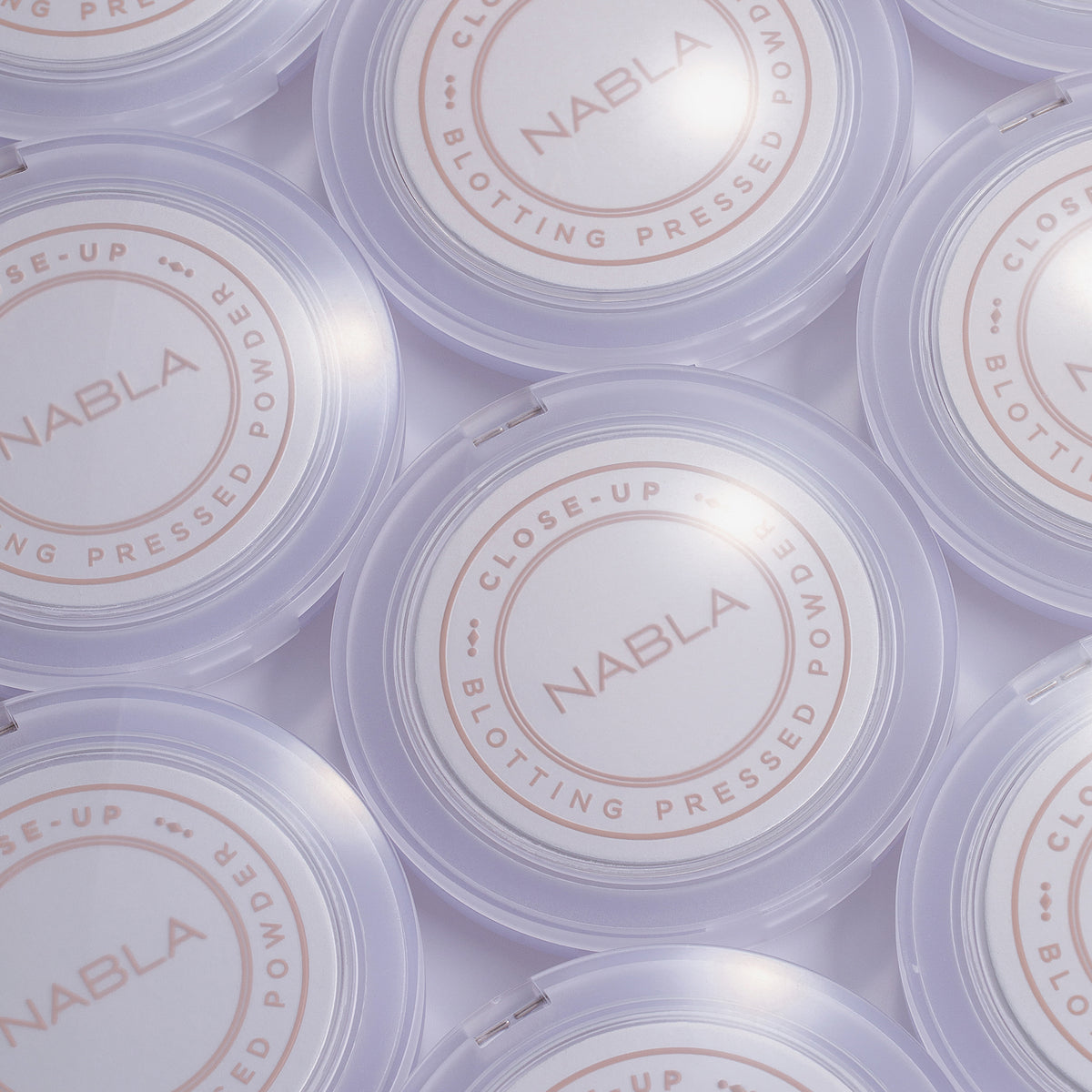 Mehrere NABLA-Puderdosen mit der Aufschrift ‚Close-Up Blotting Pressed Powder‘ in fliederfarbener Verpackung von oben fotografiert.