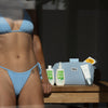 Eine Frau in einem blauen Bikini steht mit der PURISH Summer Bag 2025 – Basic Routine und einem gestreiften Beutel an einem Tisch, während das Sonnenlicht hereinströmt.