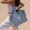Frau in weißem Outfit hält im Freien die PURISH Summer Bag 2025 – Basic Routine, eine große hellblau gestreifte Tragetasche.