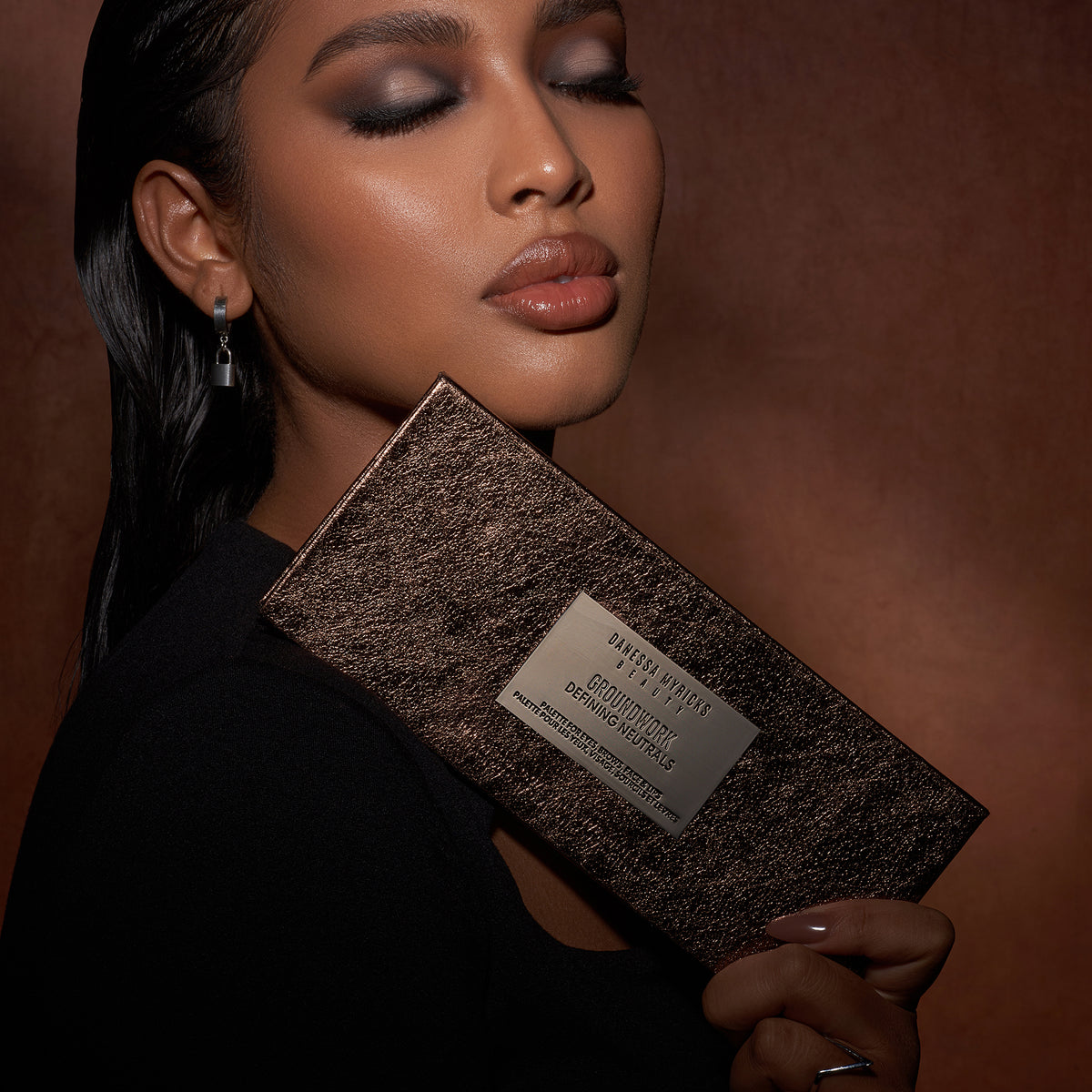 Eine Frau mit Smokey-Eye-Make-up hält die Groundwork-Palette – Defining Neutrals vor einem braunen Hintergrund.