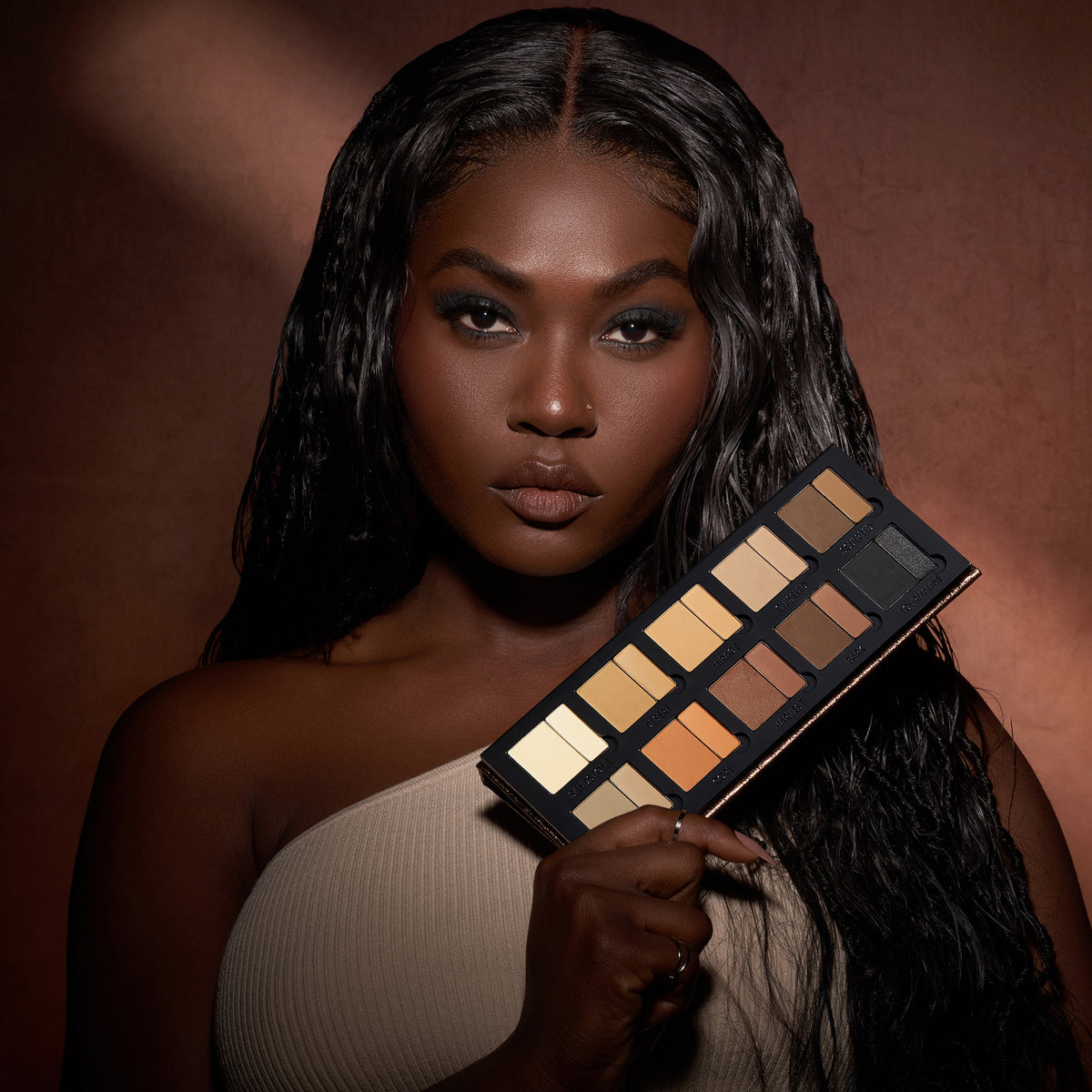 Eine Frau mit langem, dunklem Haar hält die Groundwork Palette – Defining Neutrals vor einem braunen Hintergrund.