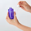 Eine Hand trägt Collagen Peptide Hydra Ampoule Serum aus einer violetten Flasche auf den Handrücken auf.