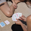Zwei Personen sitzen auf dem Boden und spielen Karten, neben ihnen liegt eine PURISH Summer Bag 2025 – Basic Routine.