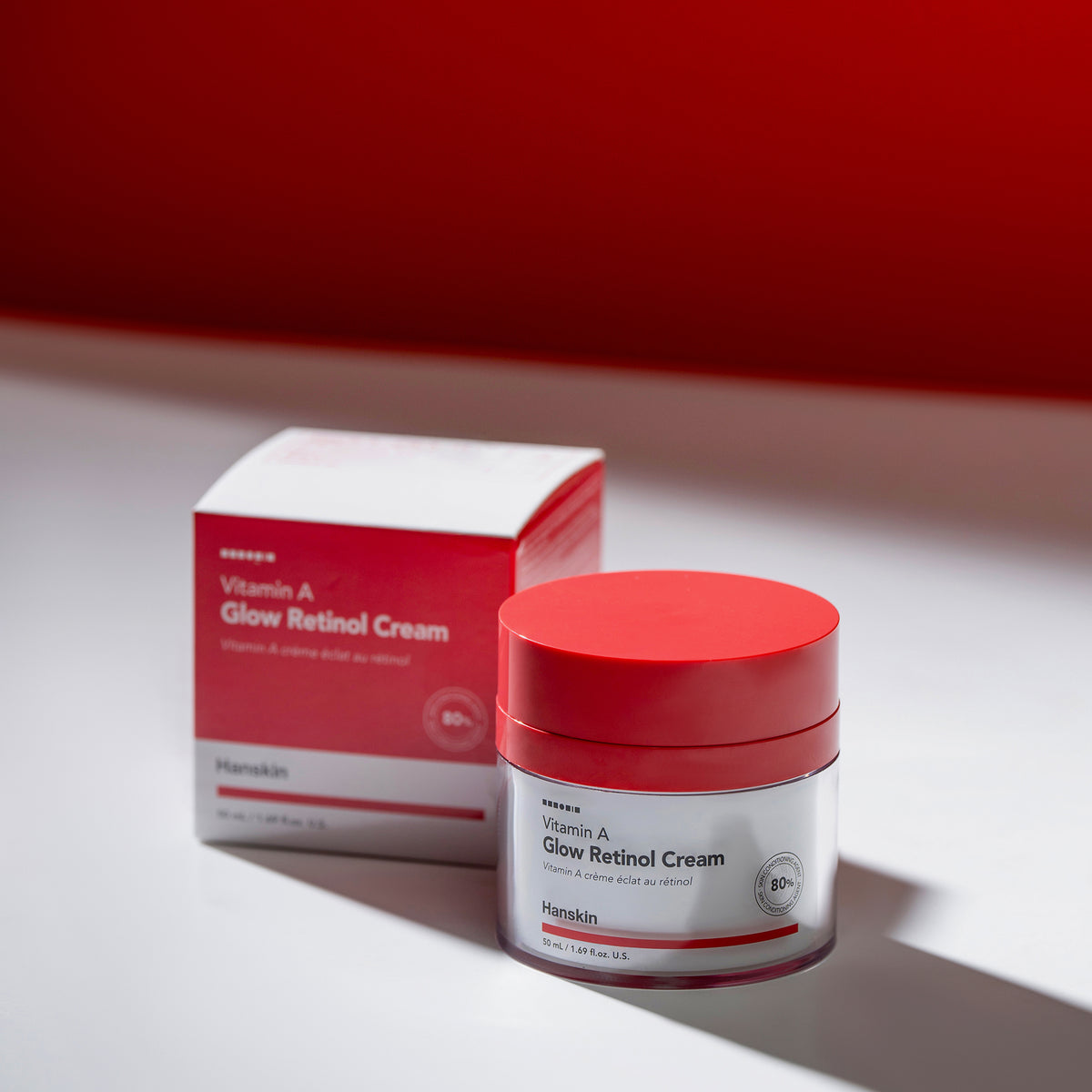 Tiegel und Schachtel mit Vitamin-A-Glow-Retinol-Creme auf einer weißen Fläche mit kräftigem roten Hintergrund.