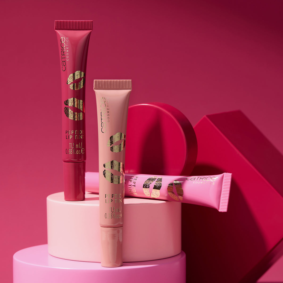 Die drei Farben des KISS & GLOW Peptide Lip Tints in Rot- und Rosatönen werden auf geometrischen Formen vor dunkelrosa Hintergrund präsentiert.