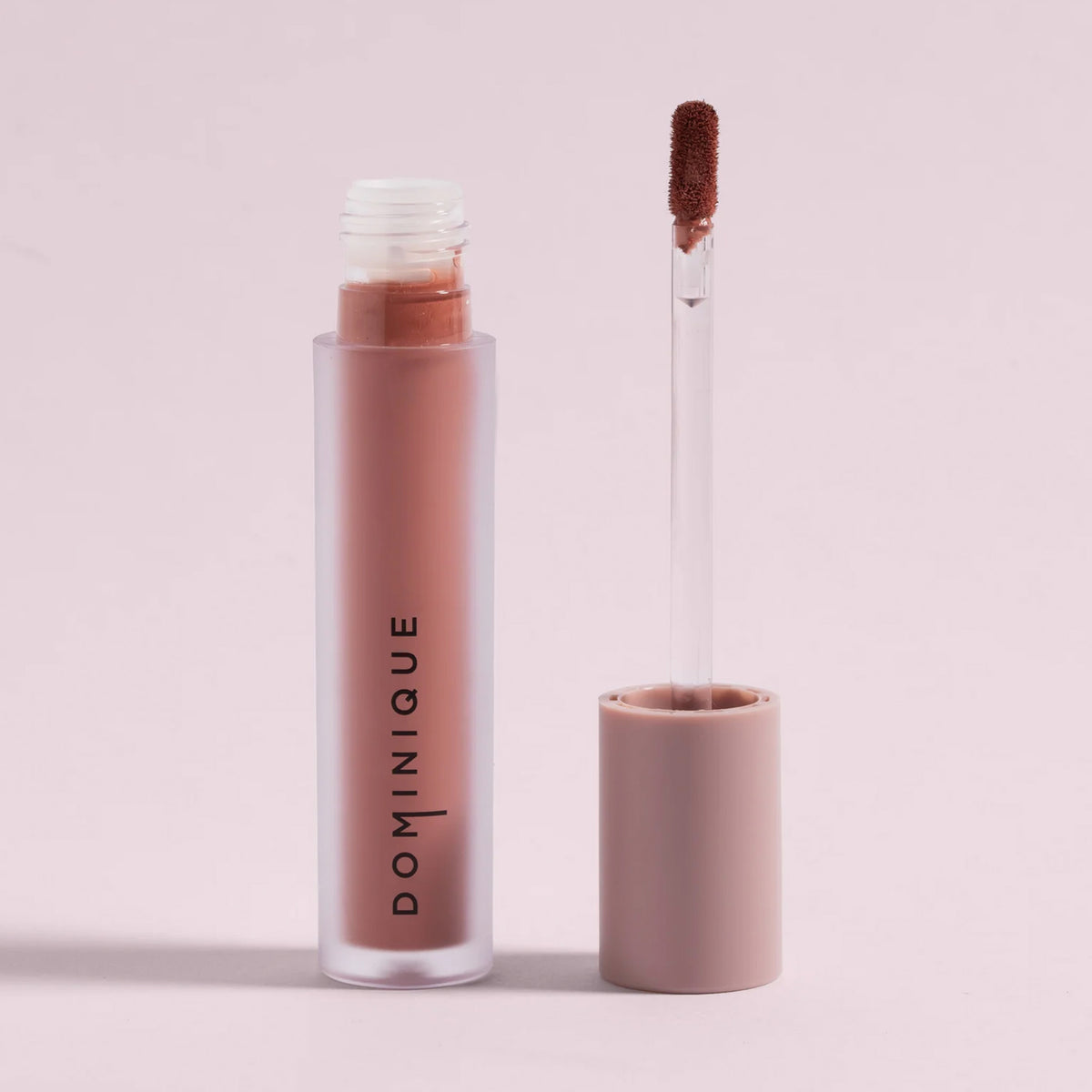 Tube mit cremefarbenem, mattem Flüssiglippenstift und Applikatorstab, beschriftet mit DOMINIQUE, auf hellrosa Hintergrund.