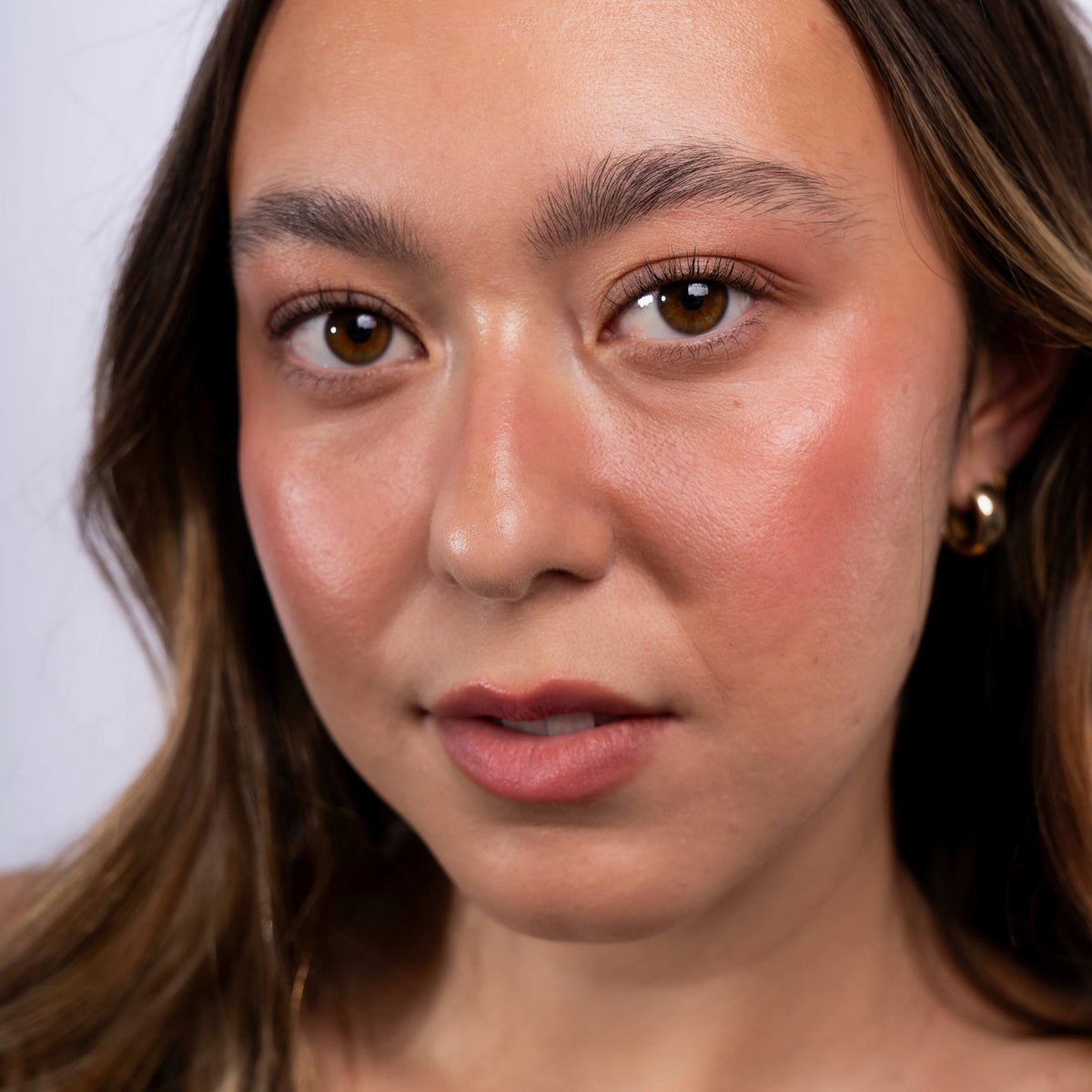 Eine Frau mit langem braunem Haar und frischem Make-up trägt Artistick Blush Sketch von LH Cosmetics und blickt vor einem schlichten Hintergrund in die Kamera.